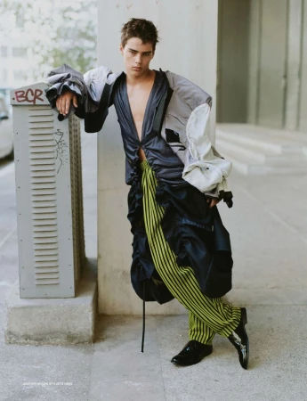 Male fashion model posing in stylish avant-garde clothing in an urban setting, leaning against a gray utility box on a city street.