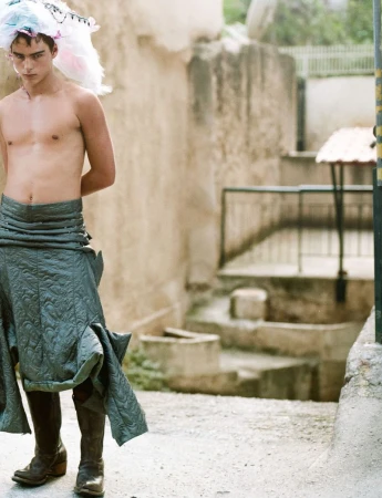 Fashion shot of a topless male model in a grey skirt and boots, posing in an alleyway with stone buildings, and wearing an eccentric hat.