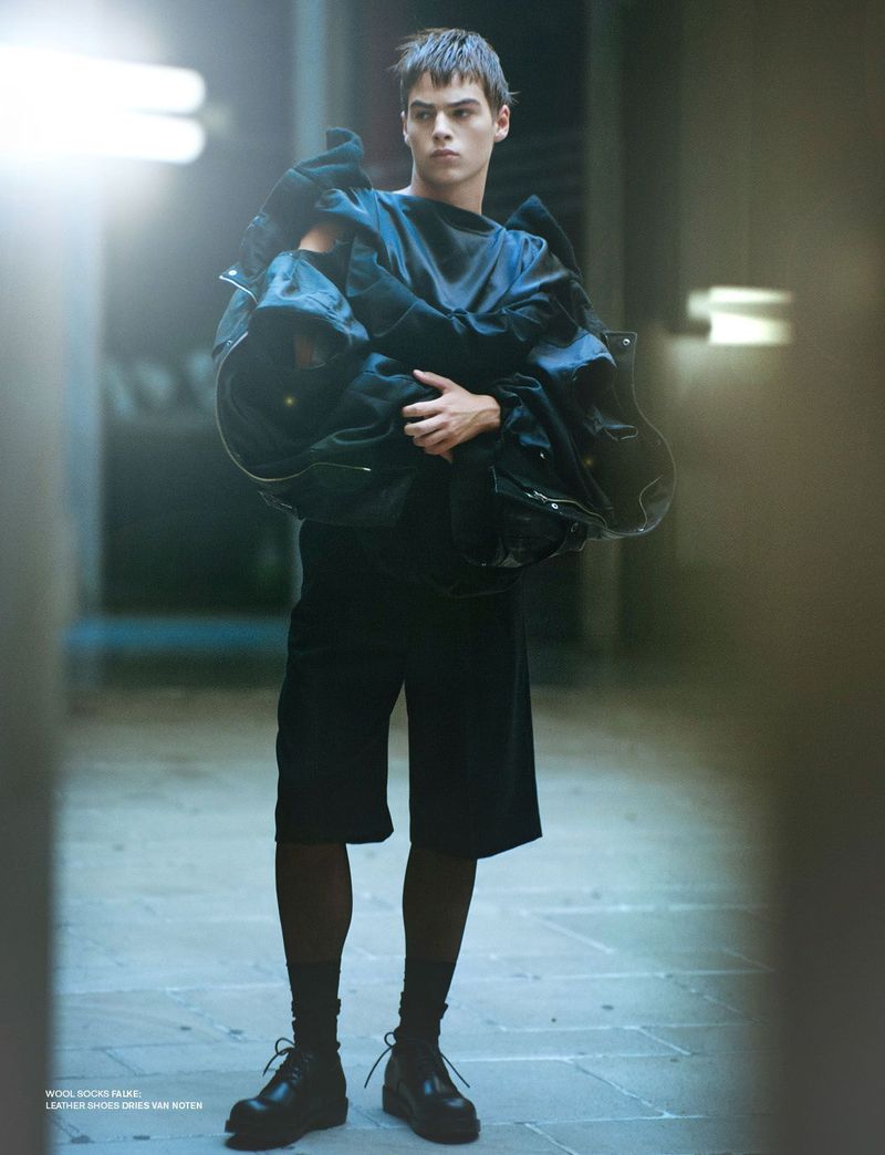 Young man standing in a hallway wearing a black outfit and holding structured garments for Dapper Dan Magazine