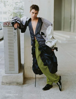 Male model leaning against a pillar wearing a deconstructed layered jacket and green striped trousers in an urban setting.