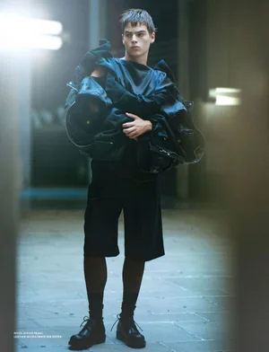 Young man standing in a hallway wearing a black outfit and holding structured garments for Dapper Dan Magazine