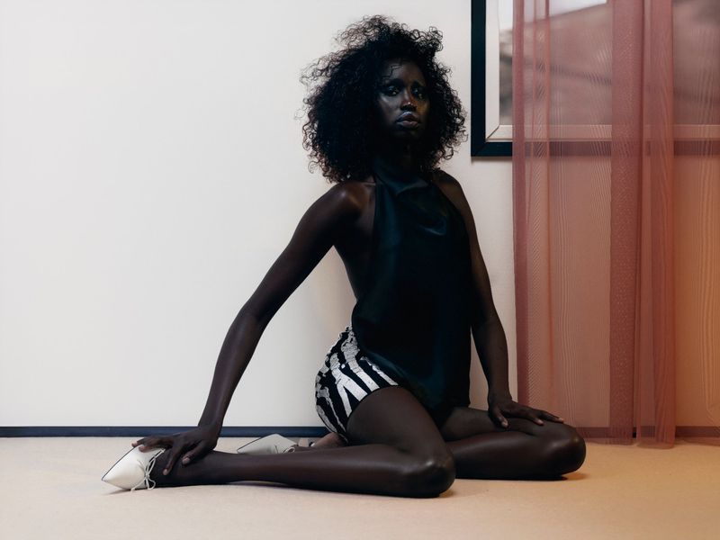 A single Black woman kneeling indoors wearing a dark top and patterned shorts, gazing forward with a neutral expression against a neutral wall.