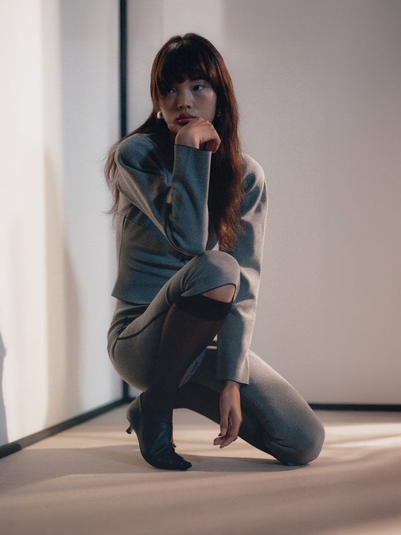 Portrait of a young Asian woman in a grey suit crouching indoors. She has long brown hair and is resting her chin on her hand.