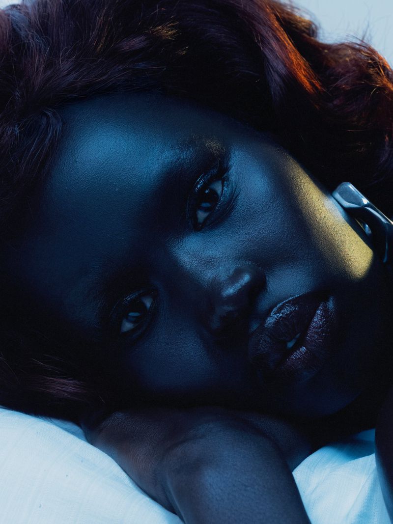 Close-up portrait of a beautiful African-American woman with dark skin, glossy lips, and red-brown hair lying on a white surface under blue lighting.