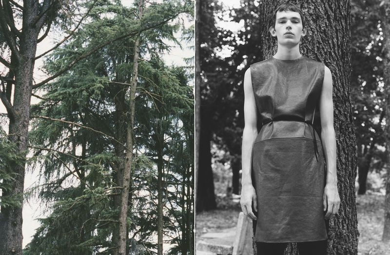 Black and white fashion shot of a slender person in a belted dress, posing outdoors near a tree, with evergreen trees in the background.