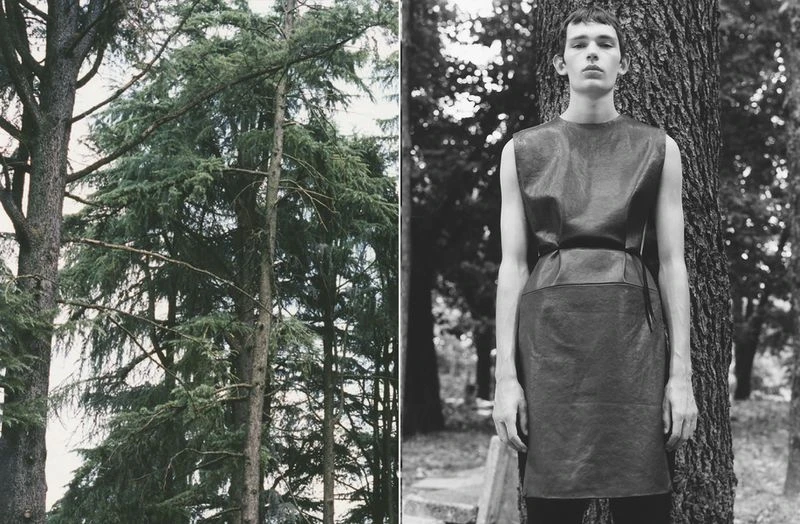 Black and white fashion shot of a slender person in a belted dress, posing outdoors near a tree, with evergreen trees in the background.