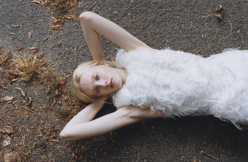 Woman in a feathered white dress lies on her back on gravel. Overhead shot captures her relaxed pose, dress texture, and neutral background.