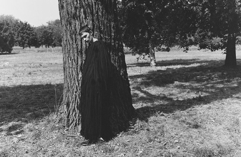 Model in a long black pleated dress leaning against a large tree trunk in a park setting.