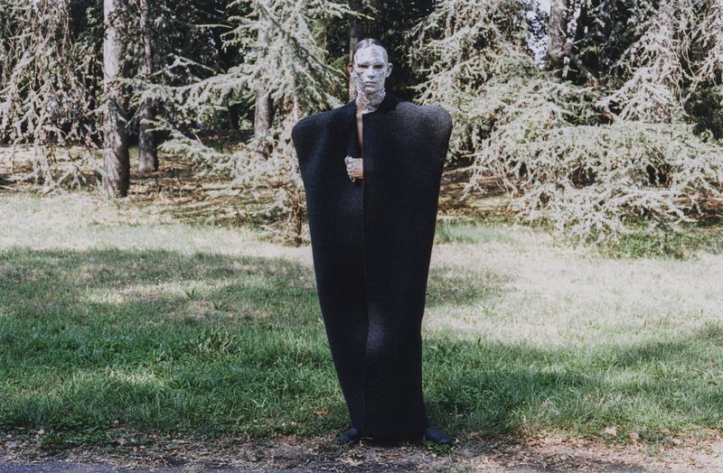 Adult posing in dramatic black cape with neutral makeup in outdoor setting surrounded by trees and grass in a fashion forward style.