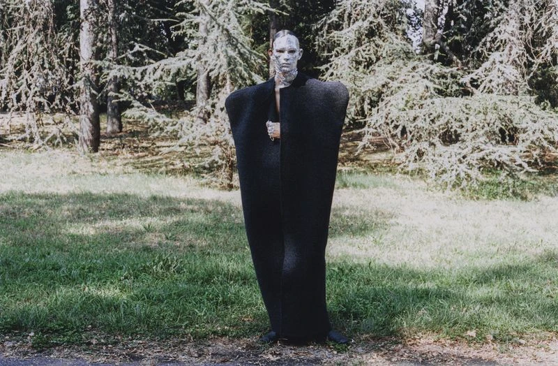 Adult posing in dramatic black cape with neutral makeup in outdoor setting surrounded by trees and grass in a fashion forward style.
