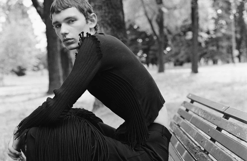 Black-and-white portrait of a male fashion model in a long-sleeved knit top sitting on a park bench, with a blurred nature background.