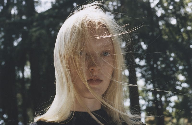 Close-up portrait of a blonde woman with hair blowing across her face in a forest setting