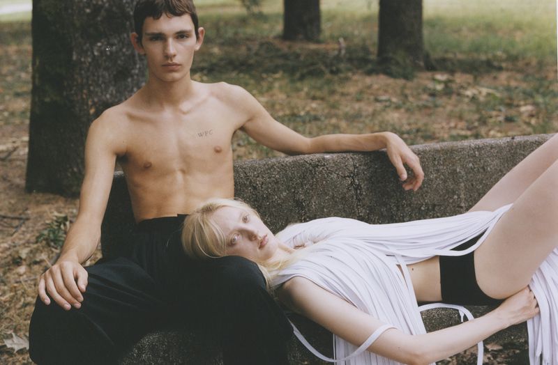 Two people pose in a relaxed outdoor setting. A shirtless man sits on a concrete bench while a blonde woman lies across his lap.