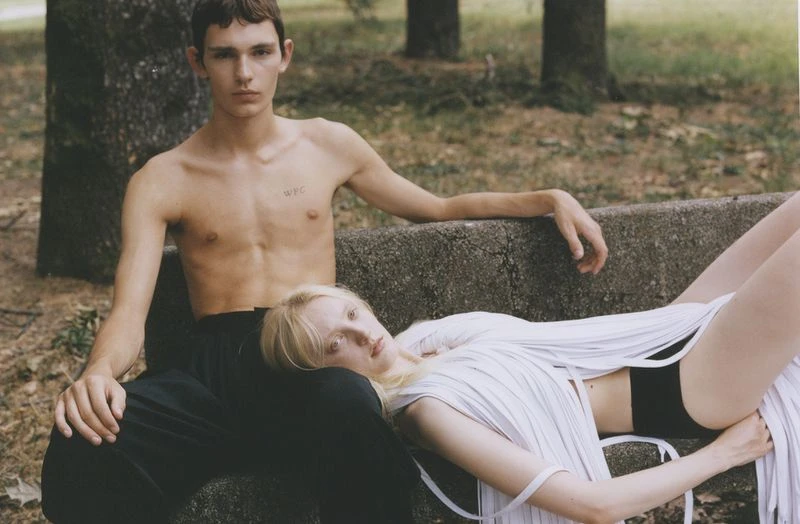 Two people pose in a relaxed outdoor setting. A shirtless man sits on a concrete bench while a blonde woman lies across his lap.