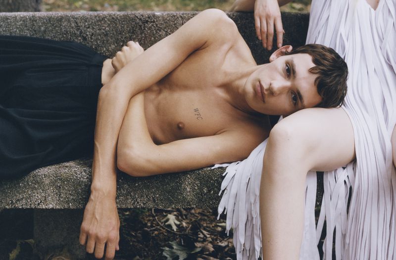 Young shirtless male model rests his head on a woman in a fringed white dress, lounging on a concrete bench in an outdoor setting.