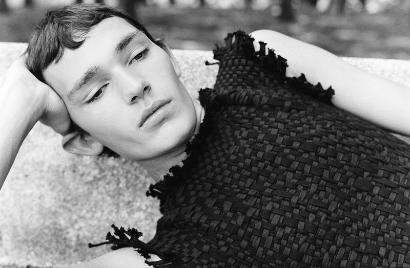 Black and white portrait of a fair-skinned young adult man lying down, wearing a textured black top, with trees blurred behind.