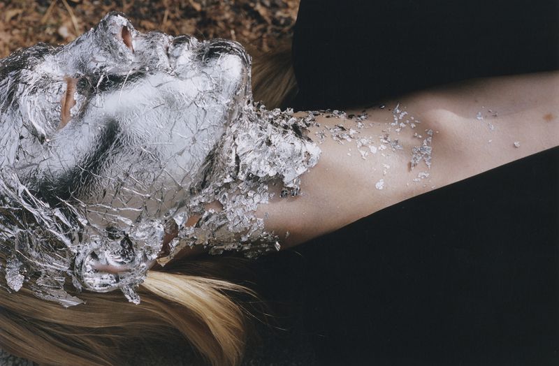 Woman's face and shoulder covered in silver foil, blonde hair visible, resting on a neutral surface against a black background.