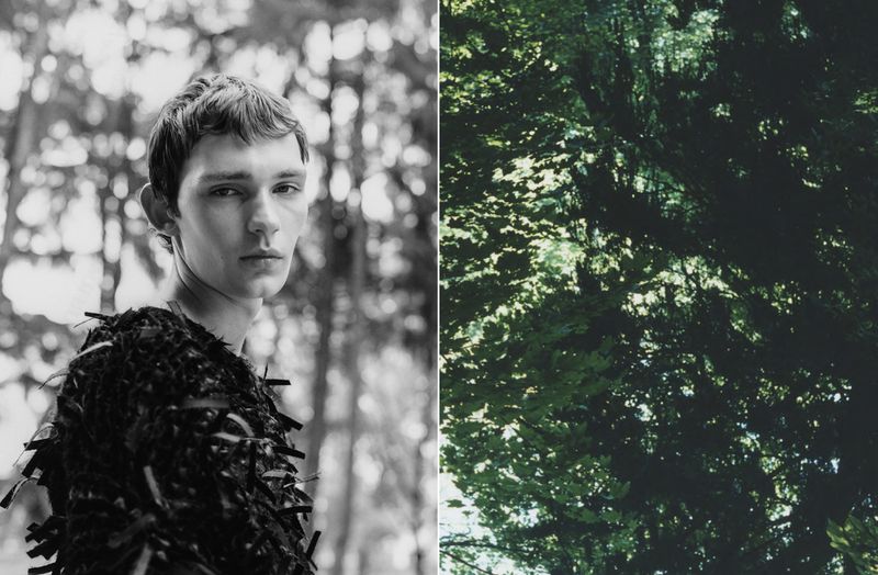 Portrait of a young man with dark hair wearing a textured top, set against a blurred forest background and foliage.