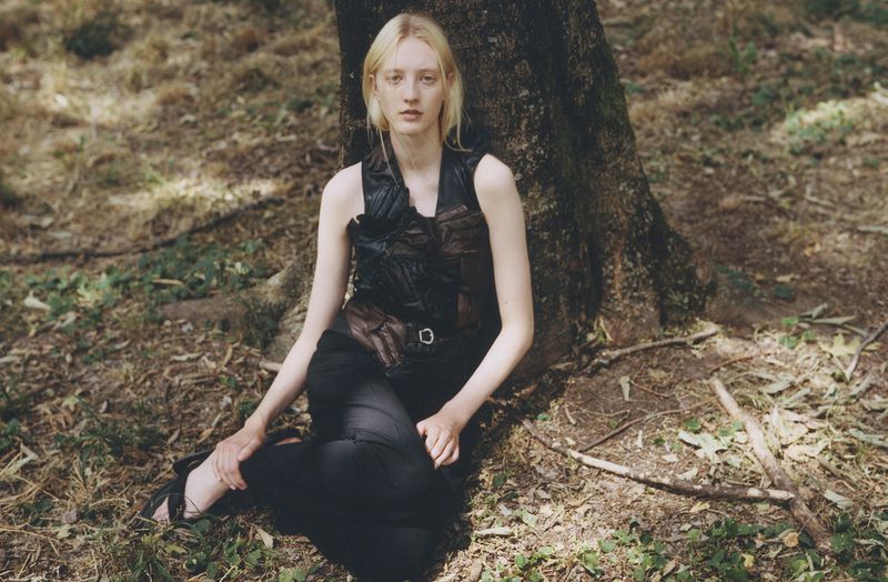 Woman in sleeveless top and dark pants sitting by tree trunk in outdoor setting with sparse ground vegetation and muted lighting.