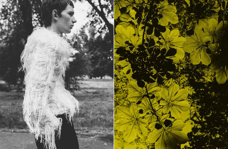 Monochrome fashion shot of a young adult male wearing an elaborate fringed top, set against a natural outdoor background with foliage.