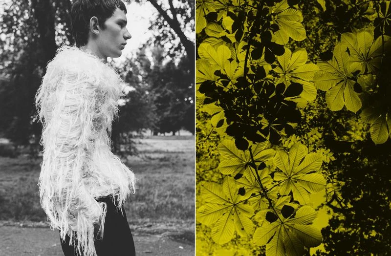 Monochrome fashion shot of a young adult male wearing an elaborate fringed top, set against a natural outdoor background with foliage.