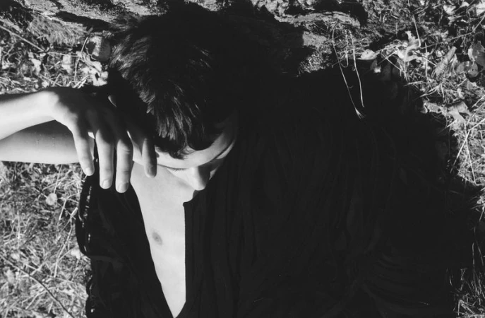 Black and white portrait of a person reclining outdoors on a grassy ground with a hand raised near their forehead.