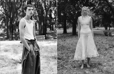 Black and white portrait of a male model in a white tank top and dark leather trousers standing outdoors in a park.