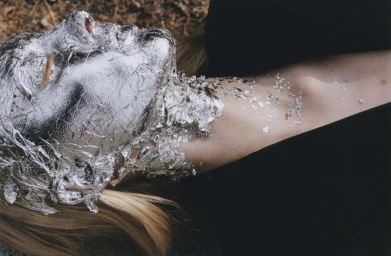 Close-up of a blonde model lying down with crinkled silver foil and metallic flakes covering her face and neck.