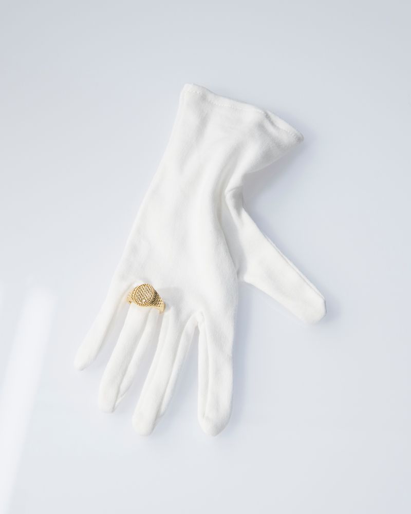 Elegant close-up of a white glove adorned with a gold ring, set against a bright white background.