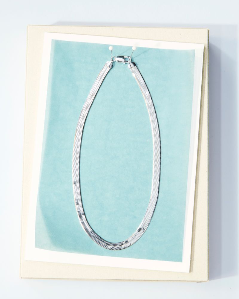 Elevated, eye-level studio shot of a flat silver herringbone necklace, pinned to a pastel blue and white background in a square frame.