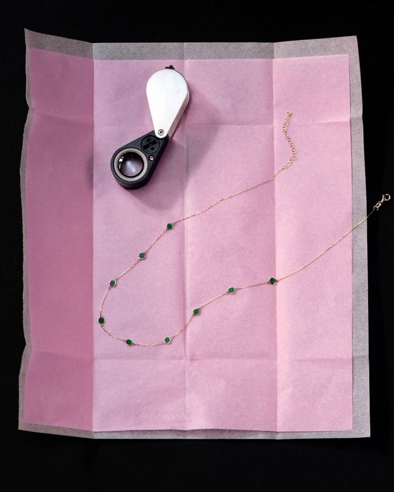 Overhead shot of a delicate gold necklace with green stones on a pink tissue, paired with a handheld magnifier for jewelry inspection.