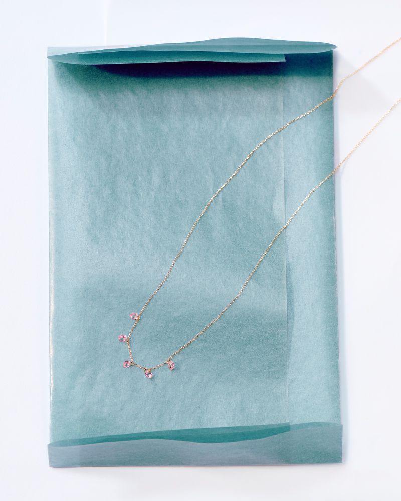 Delicate gold necklace with small pink gemstones displayed on blue tissue paper against a clean, white surface. Jewelry product shot.
