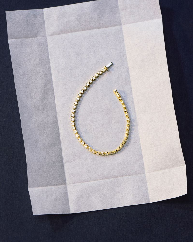 Close-up of a gold tennis bracelet with small, round diamonds displayed on a folded white tissue paper and a dark blue background.