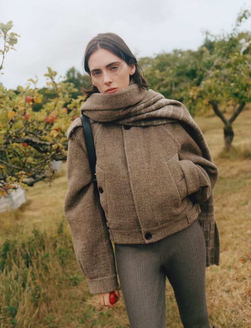 A stylish woman in a brown jacket and grey pants stands outdoors in a grassy field, showcasing a blend of fashion and nature.