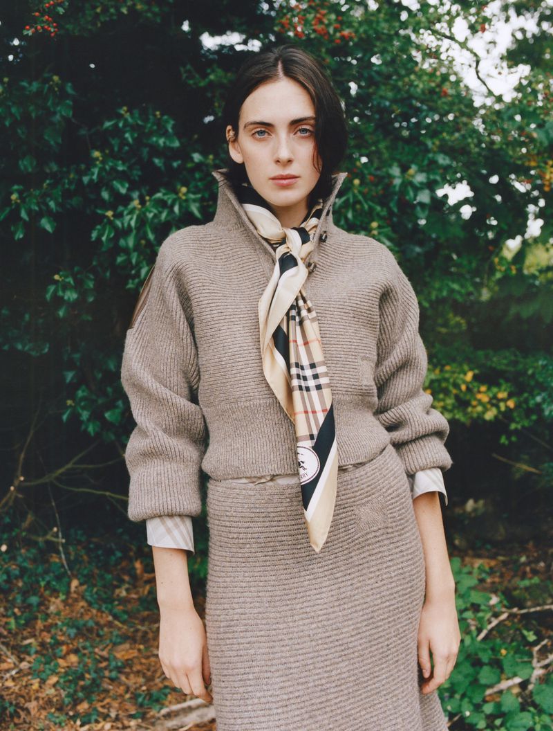 Adult woman in a beige knit outfit poses outdoors in front of a green foliage background, wearing a patterned scarf.