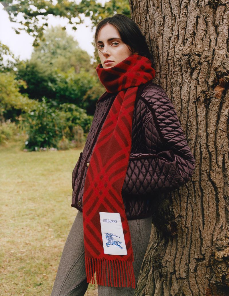 Model Lottie Morris wearing a dark quilted jacket and a red patterned Burberry scarf leaning against a large tree trunk.