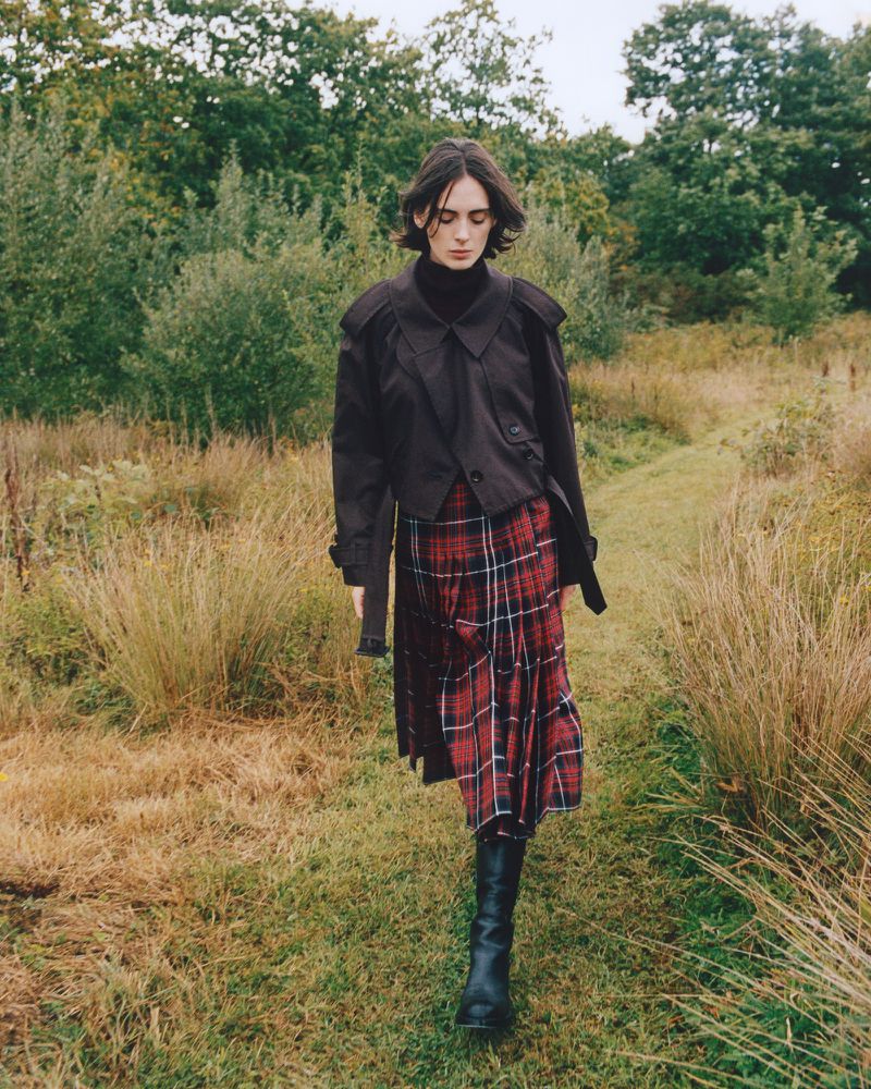 Woman walks through a grassy field wearing a black jacket, plaid skirt, and black boots, with a backdrop of trees and foliage.