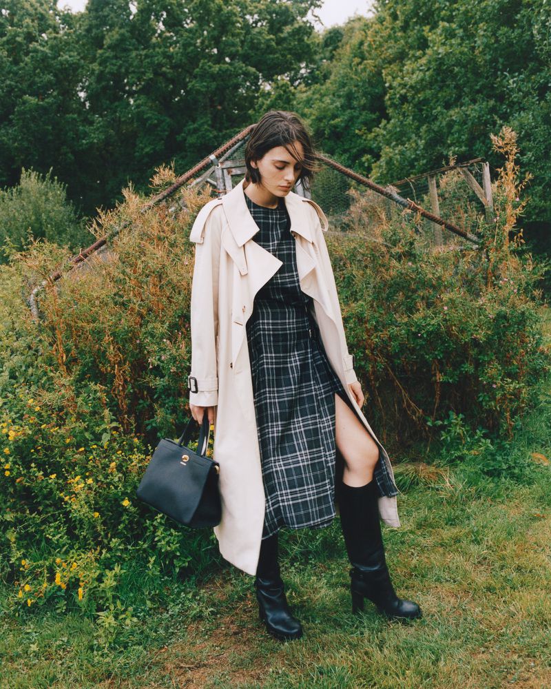 A fashionable woman in a trench coat and plaid dress stands outdoors on grass, carrying a black handbag, with trees behind.