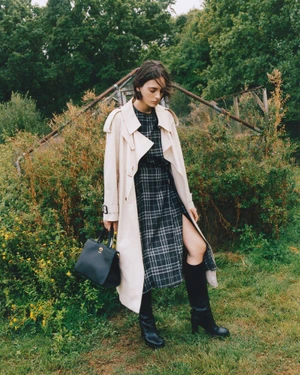 Model standing outdoors wearing a beige trench coat over a plaid dress, holding a black handbag.