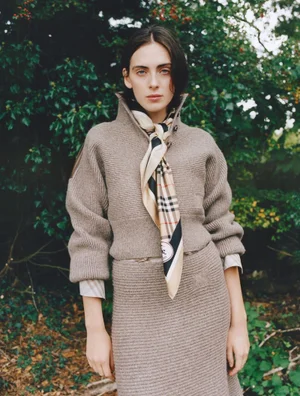 Model Lottie Morris wears a taupe ribbed knit sweater, matching skirt, and signature check silk scarf outdoors against green foliage.