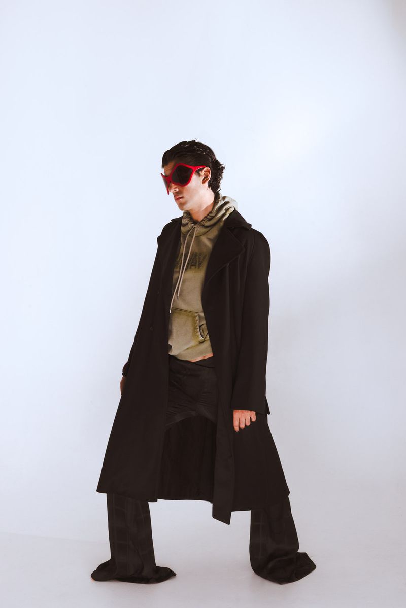 Adult male poses in a studio wearing red sunglasses, a black coat, a khaki-colored hoodie, and black pants against a white background.
