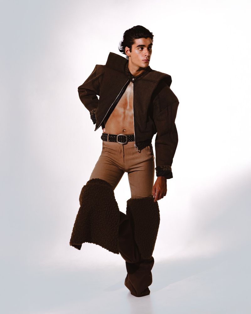 Fashionable adult male posing in a cropped brown jacket, tan pants, and brown leg coverings against a white background in a studio.