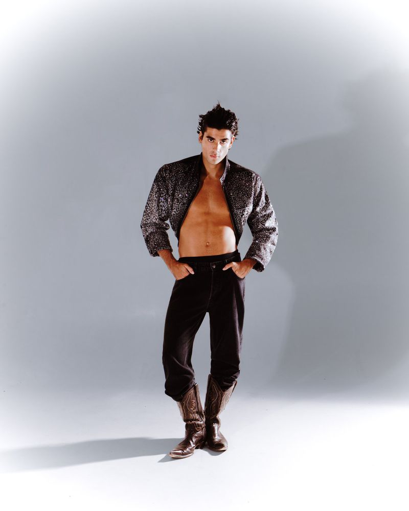 Full-length studio portrait of an adult male wearing a bolero-style jacket, dark denim, and cowboy boots against a light grey backdrop.