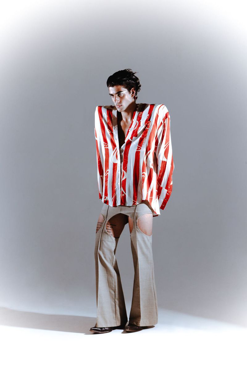 Fashion-forward young adult male posing in red and white striped shirt and beige cutout pants against a neutral studio backdrop.
