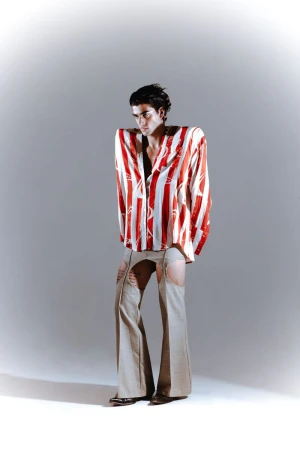 Fashion-forward young adult male posing in red and white striped shirt and beige cutout pants against a neutral studio backdrop.