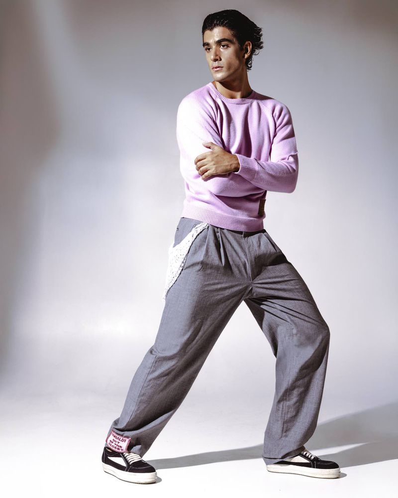 Young man in a pale pink sweater and grey trousers posing against a grey gradient studio background