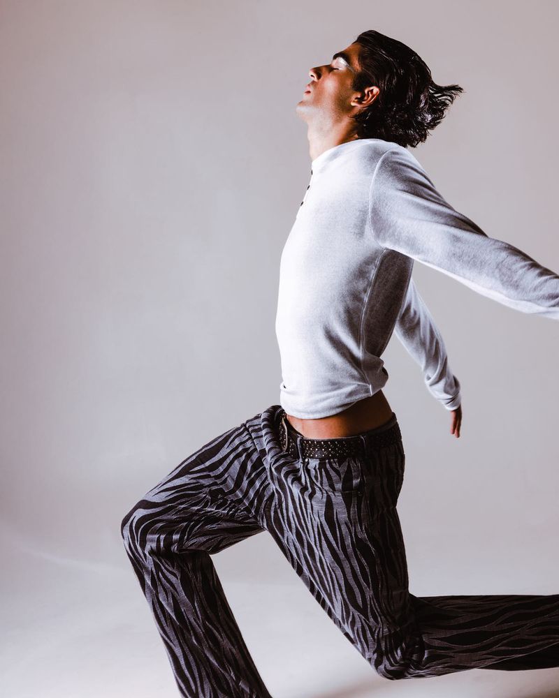 Man in zebra-striped pants and a long-sleeve white shirt lunges forward with arms outstretched in a light-filled, neutral studio.