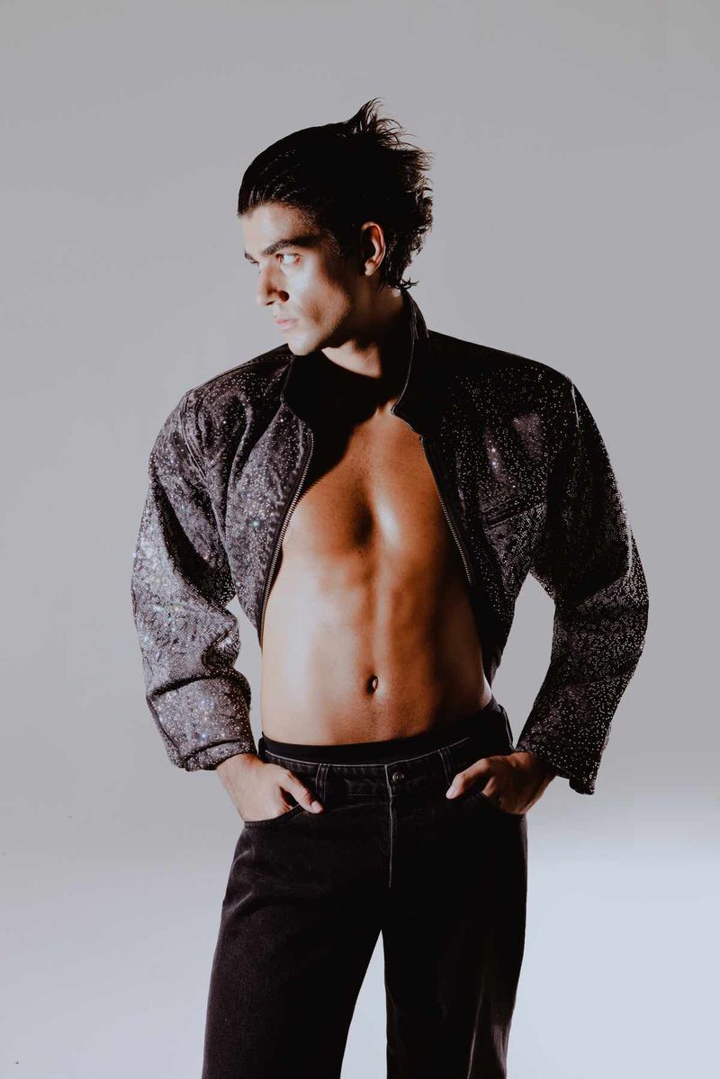 Man in stylish bolero jacket and jeans strikes a pose against a plain background, showcasing fashion and confidence.