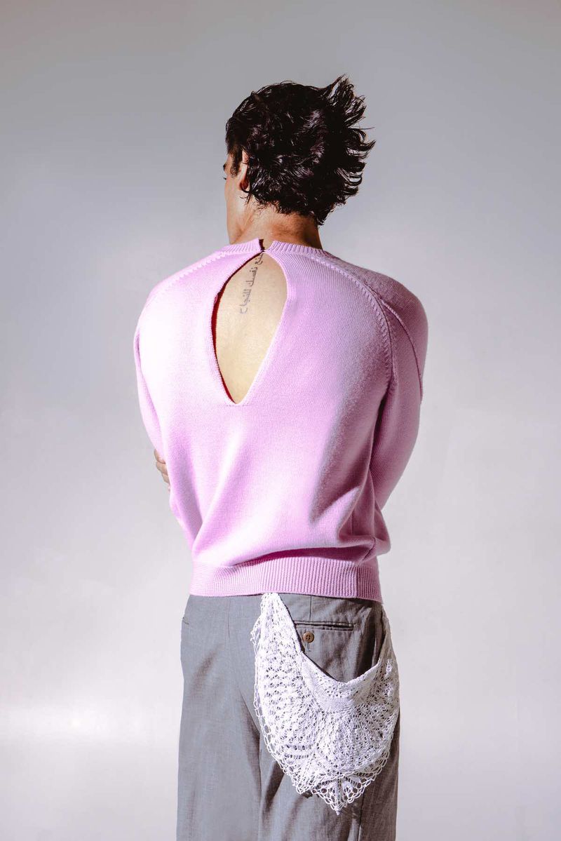 Back view of adult male with dark hair in a pink sweater and gray pants, featuring a lace pocket square against a plain studio backdrop.