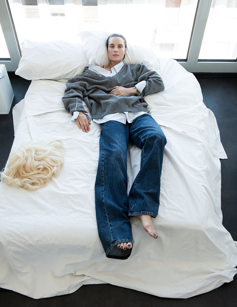 Model Charlotte Tomas lies on a white bed in a grey knit sweater and blue jeans, with a blonde wig beside her.